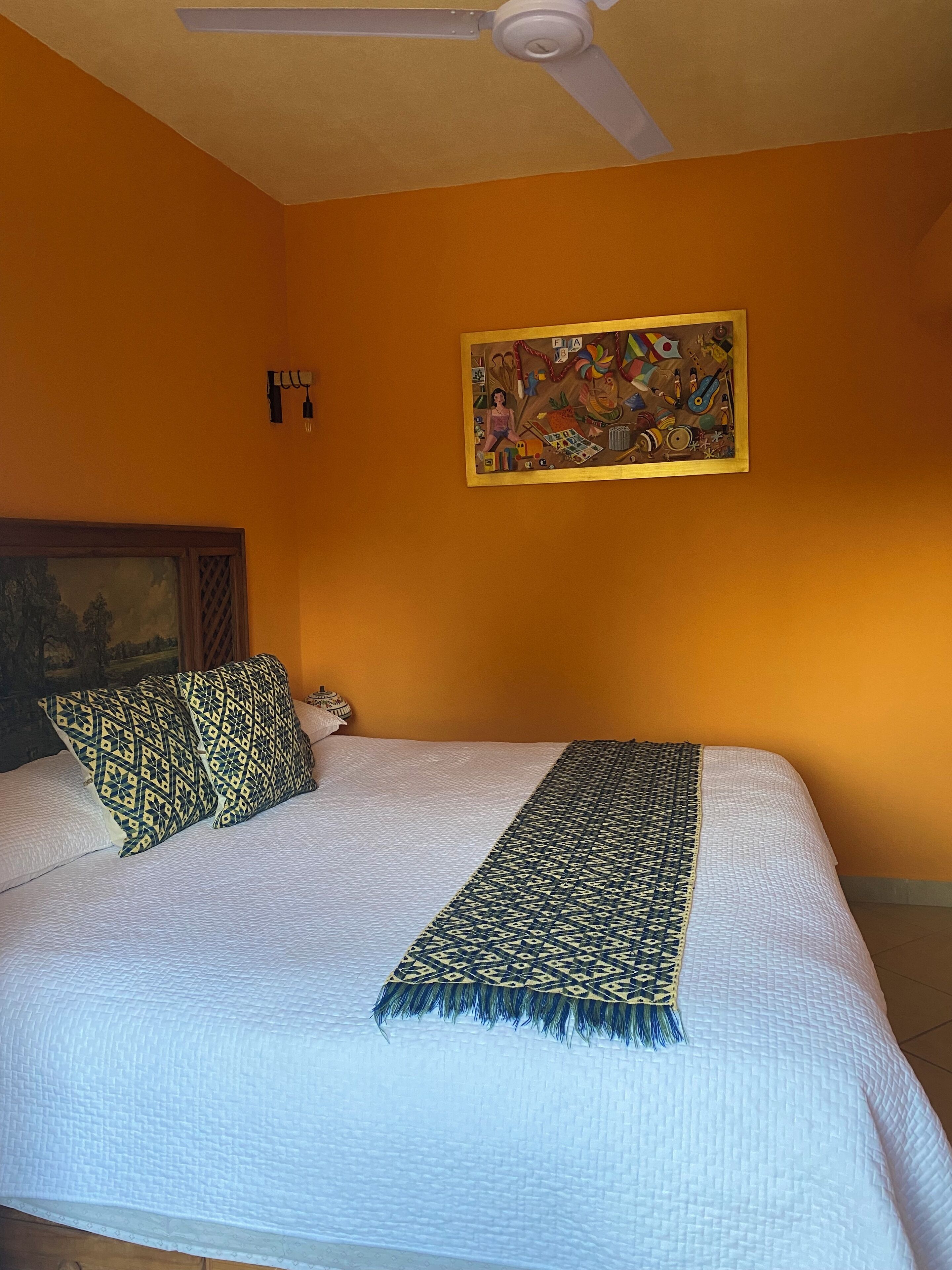 Une chambre d’hôtel avec un lit, des oreillers, une couverture, un ventilateur de plafond et un tableau au mur.