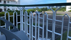 A balcony with a white metal railing overlooking a street with parked cars and a building with a red roof.