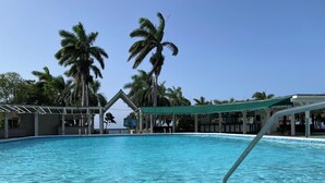 A clear blue pool with a tiled edge, surrounded by palm trees and a covered area with a green roof.