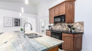 Una cucina con piano di lavoro in granito, lavello in acciaio inox, pensili in legno, forno a microonde e schienale piastrellato.