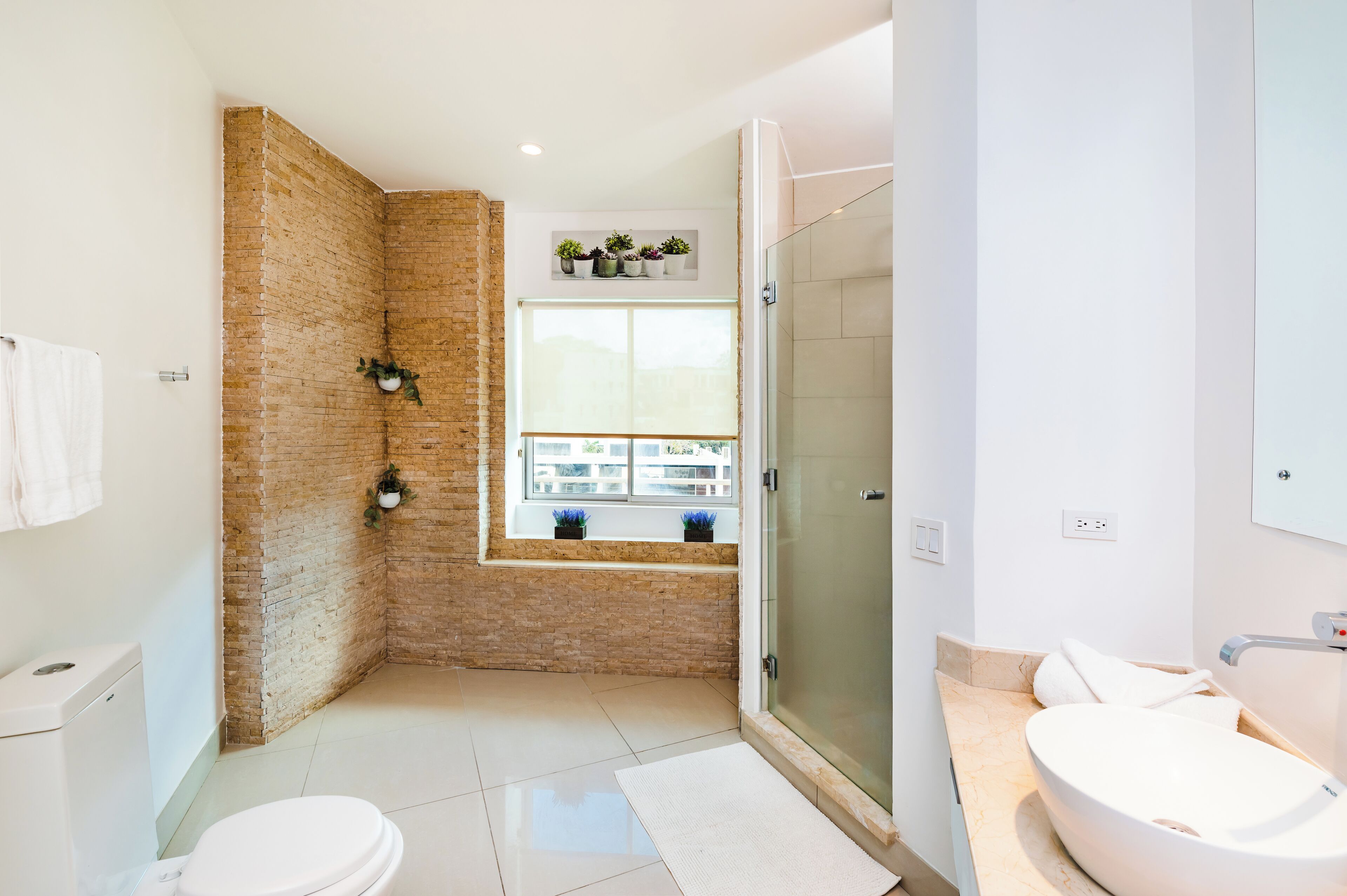 A modern bathroom with a brick accent wall, a glass shower enclosure, a white sink, and a window with potted plants.