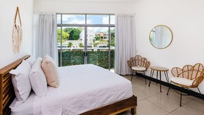 Une chambre avec un grand lit, deux fauteuils, une petite table et une vue sur la verdure et les bâtiments environnants.