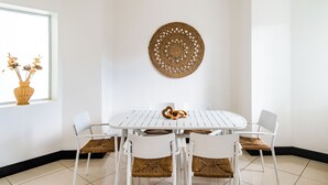 Un coin repas avec une table et des chaises blanches, une décoration murale en fibres tressées et un vase en osier contenant des fleurs séchées, posé sur un rebord de fenêtre.
