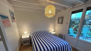 A bedroom with a striped bedspread, two bedside lamps, a wicker pendant light, and a view of outdoor chairs through a window.