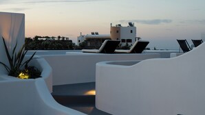 A rooftop terrace with white walls and curved seating areas, overlooking a coastal landscape. - Griseo Villas (Santorini)
