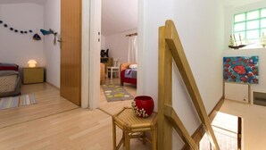 Habitació amb terra de fusta, un llit amb llençol vermell, una taula petita amb un vas vermell i un quadre a la paret.