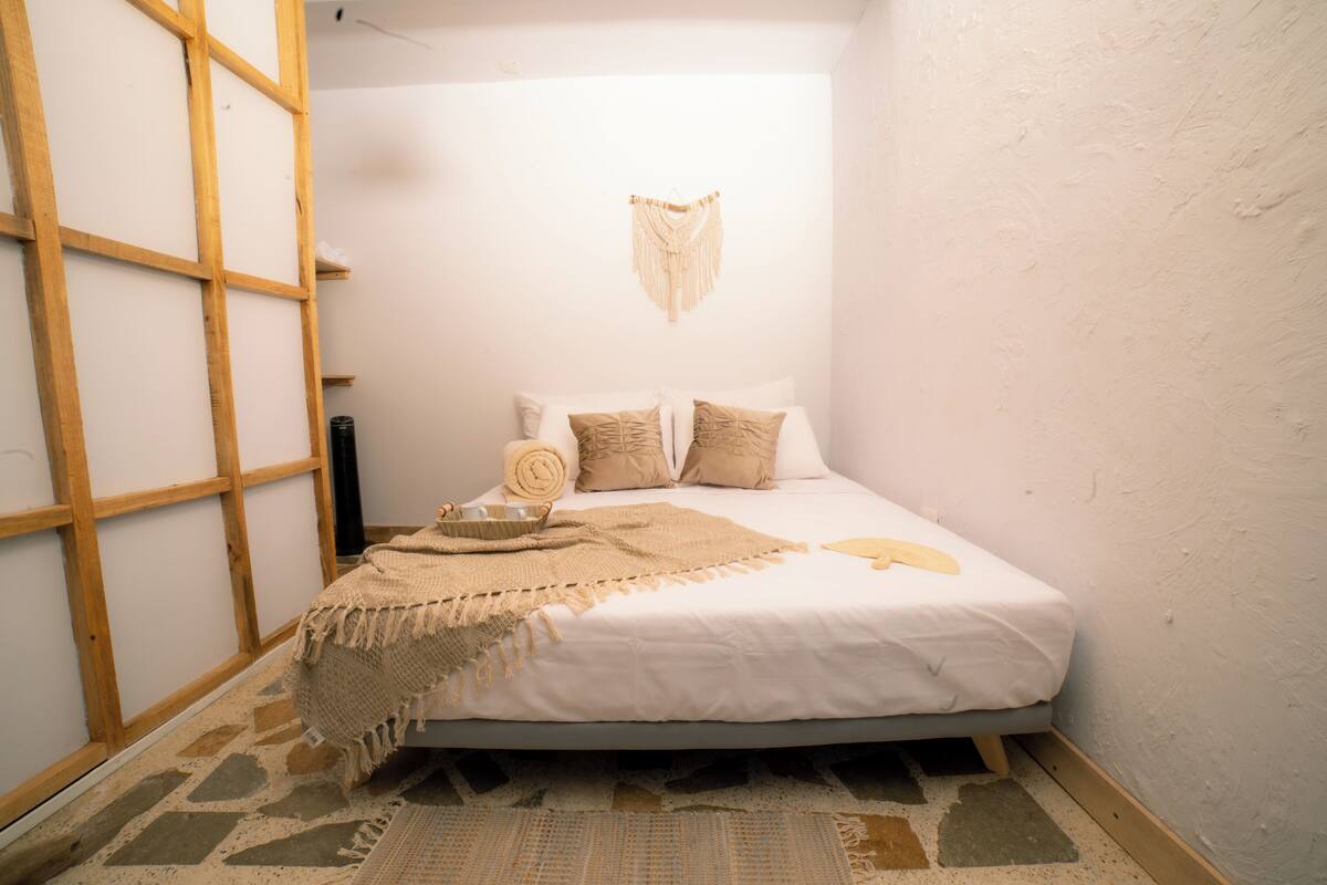 A bedroom with a bed, a wooden ladder, a wall-mounted macrame piece, and a patterned rug.