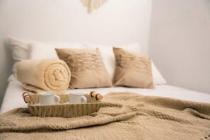 Cama bem arrumada com um cobertor bege, duas toalhas enroladas e uma bandeja com duas canecas brancas e um apoio de cortiça.