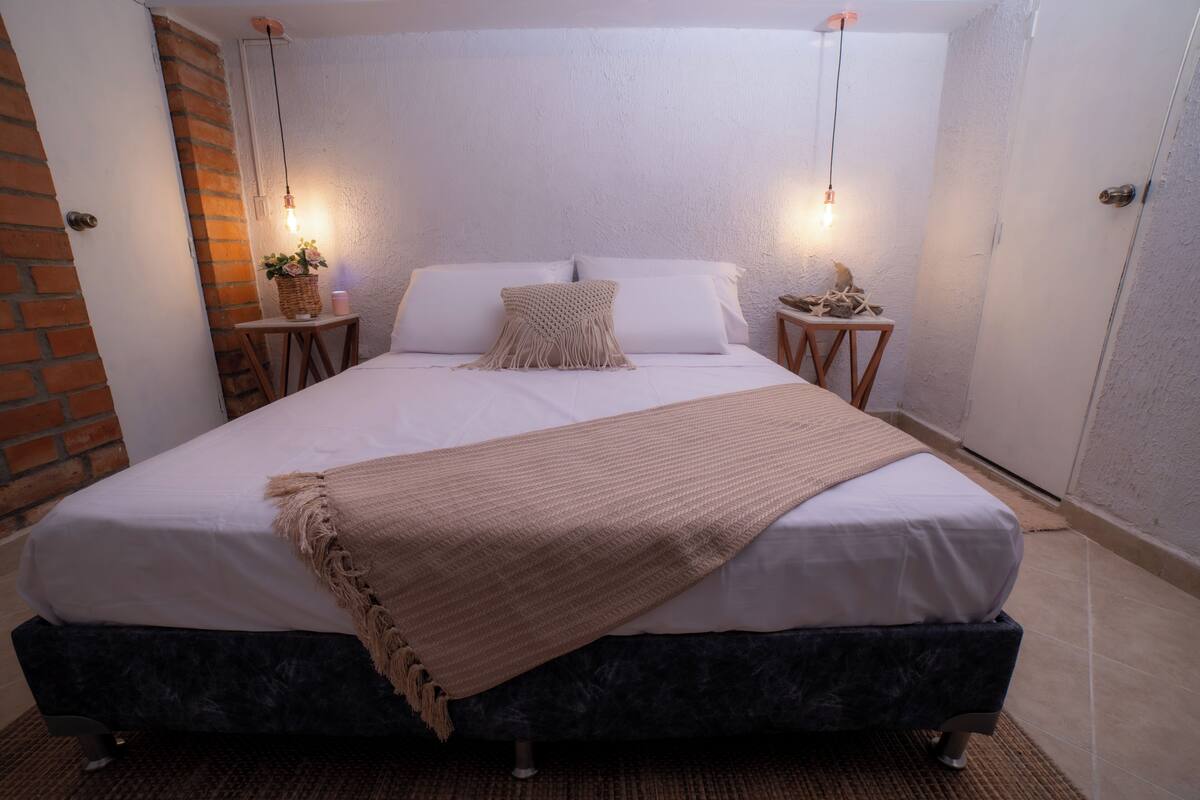 A neatly made bed with a beige throw blanket, two bedside tables with decorative items, and a brick wall adjacent to the room.