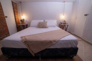 Uma cama bem arrumada com um cobertor bege, duas mesas de cabeceira com objetos decorativos e uma parede de tijolos adjacente ao quarto.