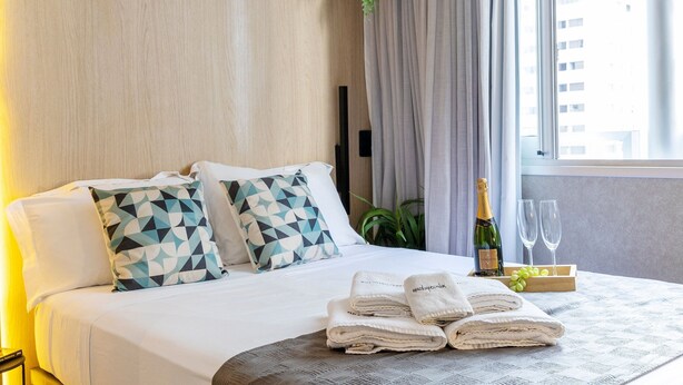 Una camera d'albergo con un letto rifatto, due cuscini a motivi geometrici, una bottiglia di champagne, due bicchieri e un vassoio con grappoli d'uva.