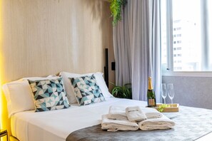 A hotel room with a neatly made bed, two geometric patterned pillows, a bottle of champagne, two glasses, and a tray with grapes. - Apartamento moderno com piscina e AC (São Paulo)