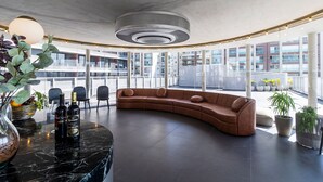 Zona relax moderna con un divano marrone curvo, un tavolino in marmo con due bottiglie e un'ampia pianta in vaso.