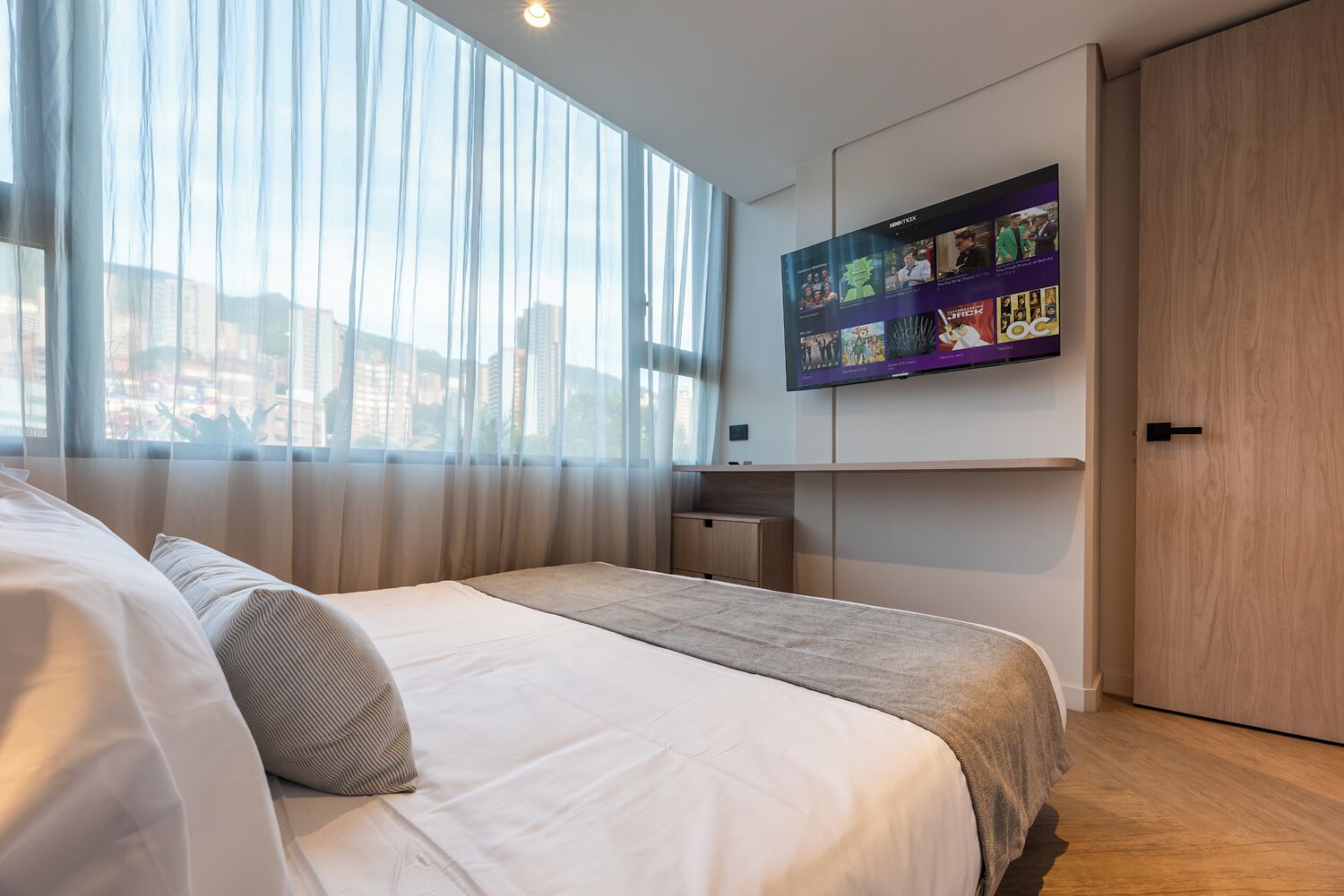 Un dormitorio moderno con una cama grande, un televisor montado en la pared y vistas a la ciudad a través de cortinas translúcidas.