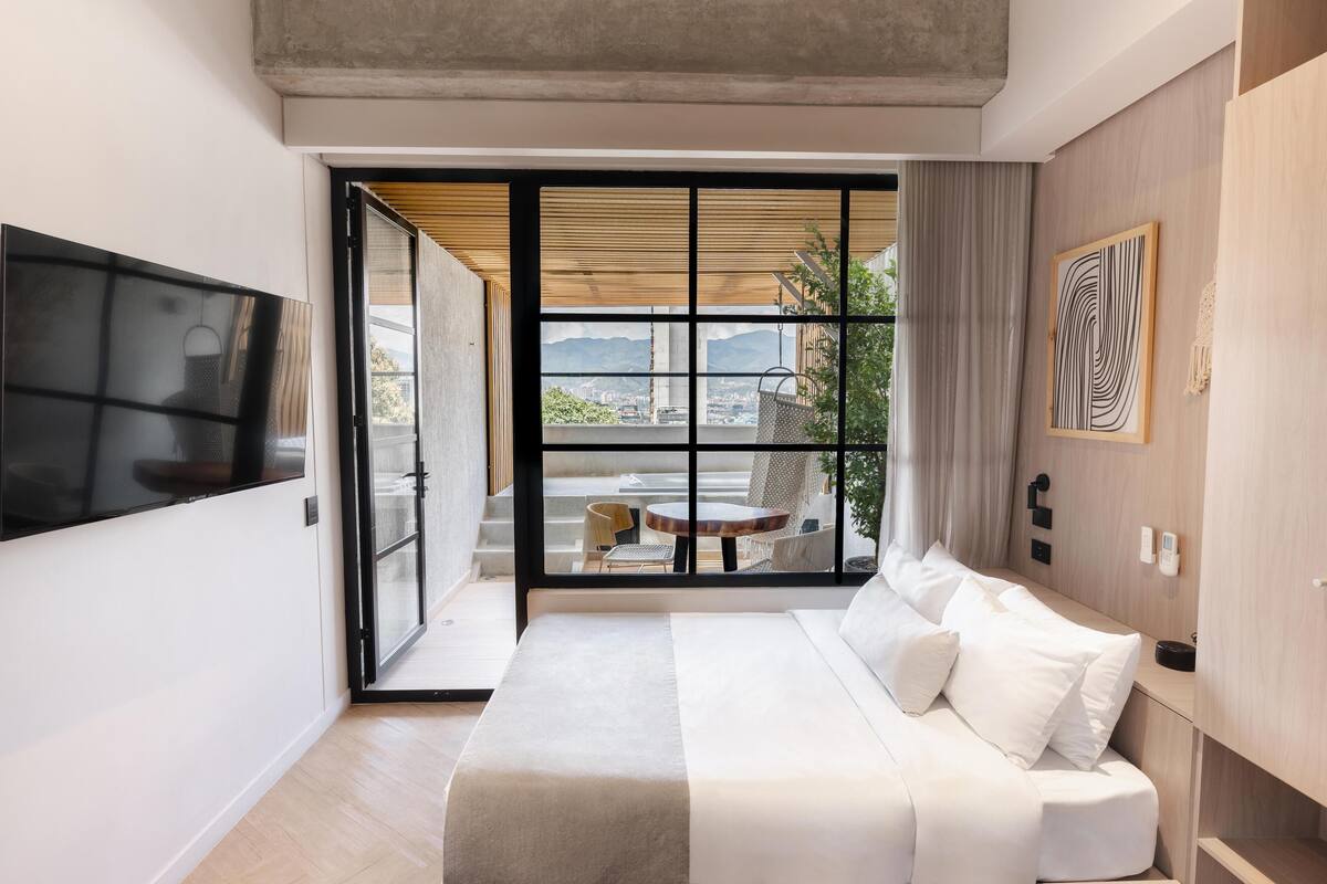 Ein modernes Hotelzimmer mit Bett, Fernseher, einer Schiebetür zu einem Balkon mit Tisch und Stühlen sowie Blick auf die Stadt.