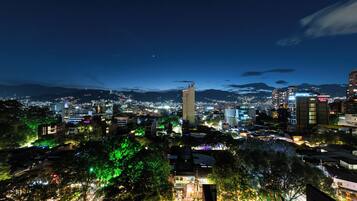 Un paisaje urbano nocturno con edificios iluminados, una torre destacada y vistas a montañas distantes.