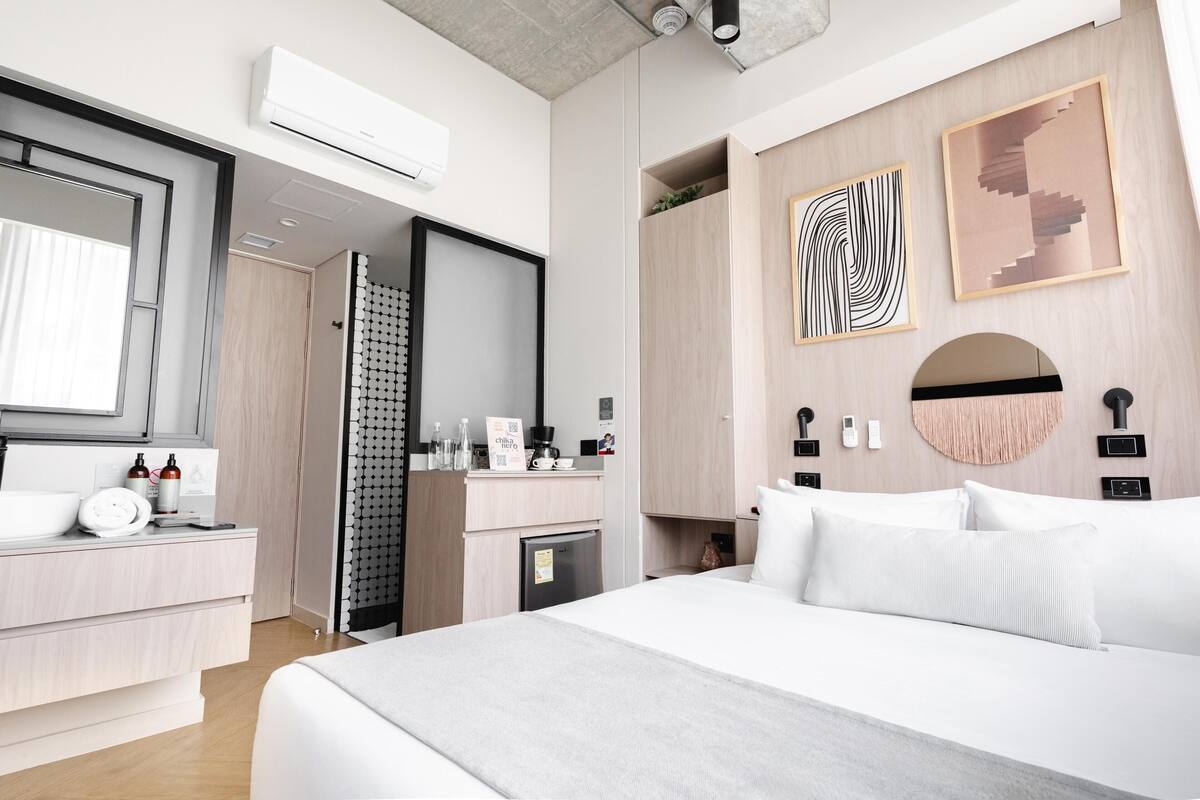 Ein modernes Hotelzimmer mit Bett, Schminktisch mit Spiegel, einer kleinen Küchenzeile und Bildern an den Wänden.
