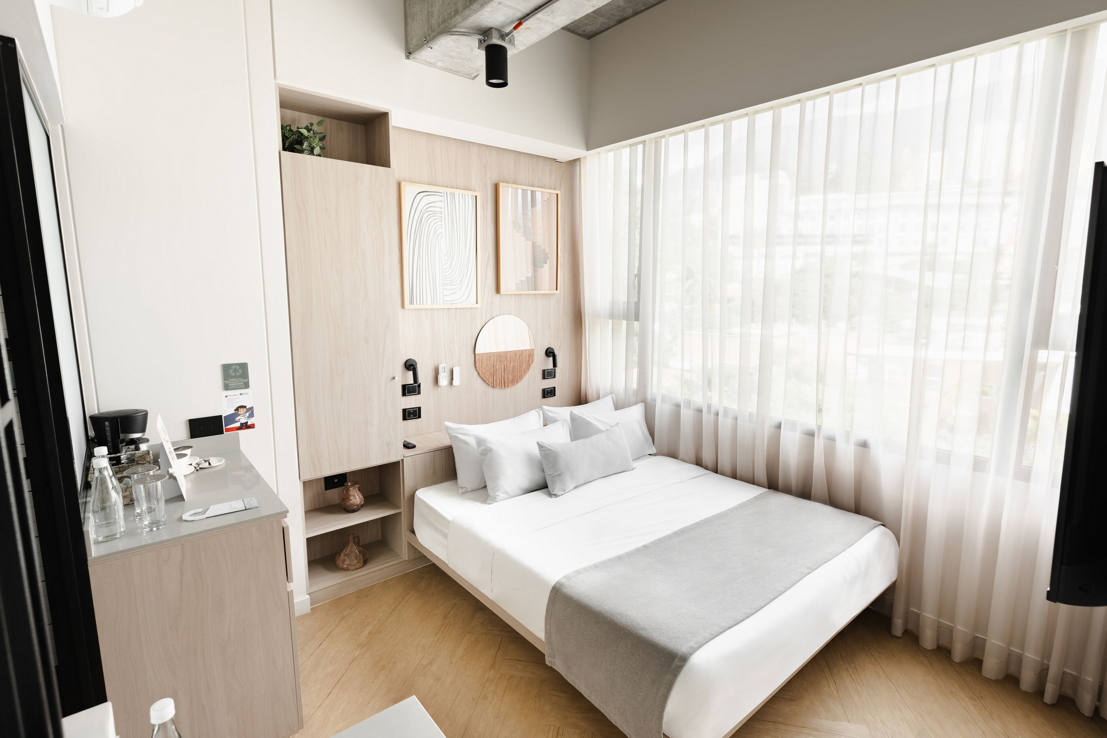 A modern hotel room with a large bed, a desk with a chair, and a window with sheer curtains.
