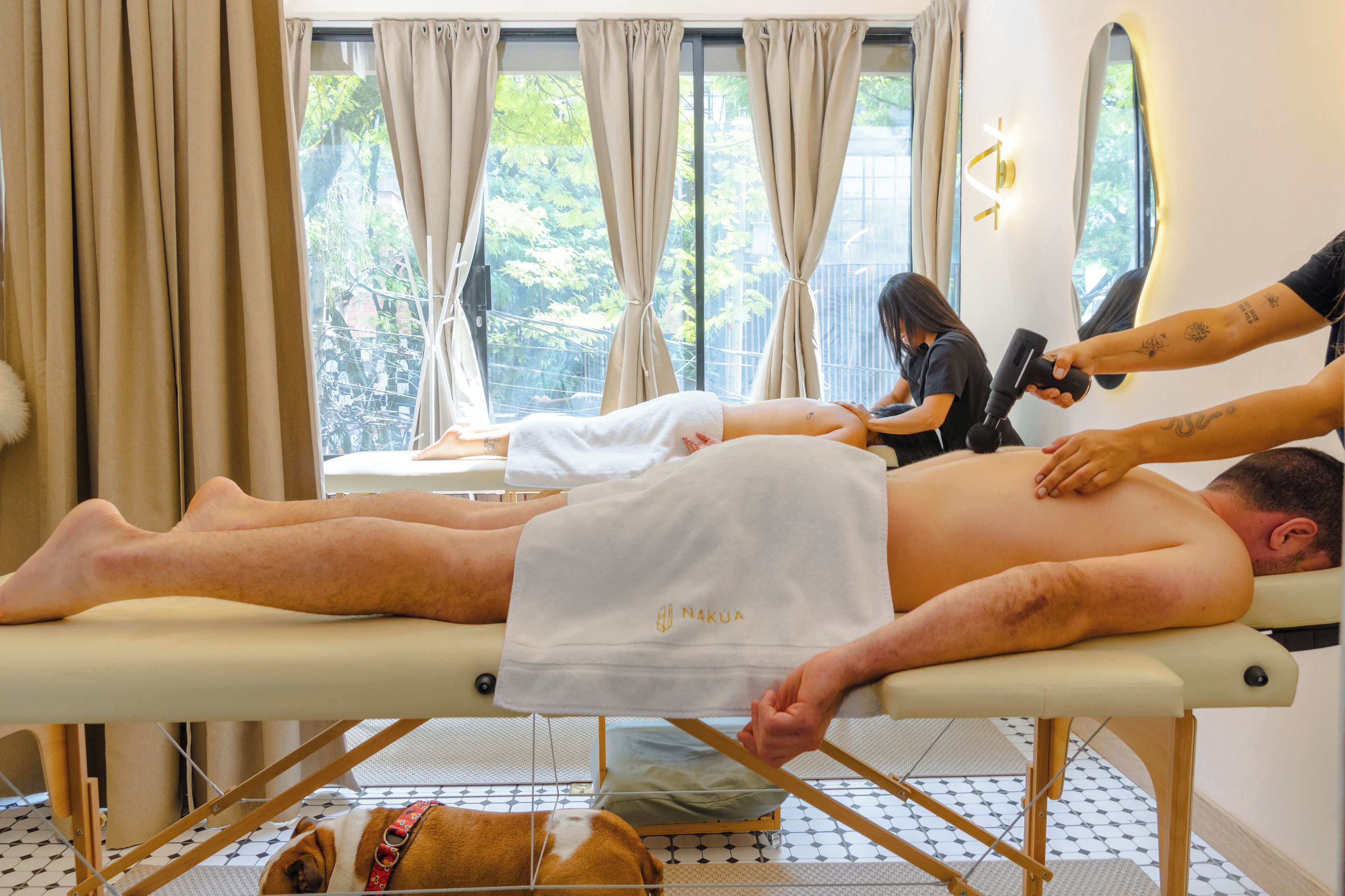 A massage therapy room with a client lying on a treatment table, a therapist attending to the client, and a dog resting on the floor.
