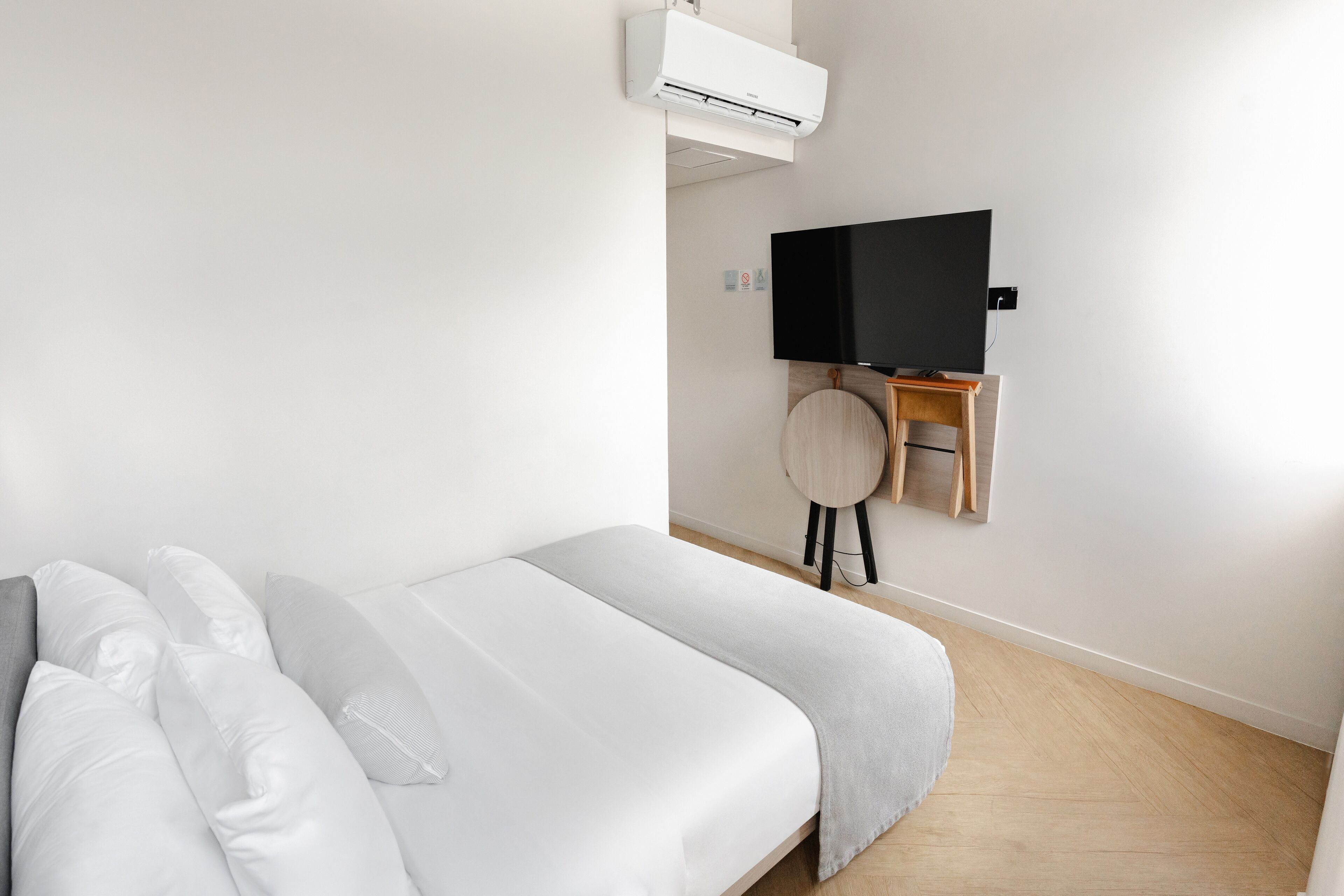 A minimalist hotel room with a bed, a mounted TV, and a small wooden side table.