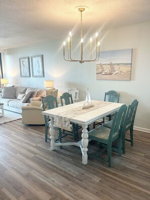 A dining area with a white table, green chairs, and a chandelier. There is a sofa and a painting on the wall.