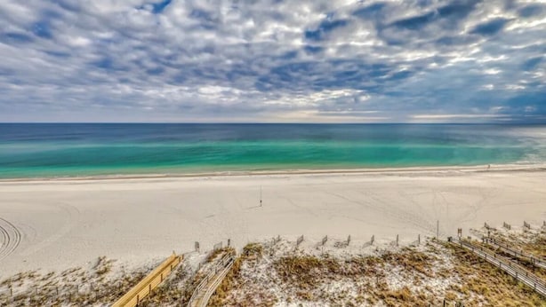 A beach with a wooden boardwalk leading to the shore, clear turquoise water, and a cloudy sky.