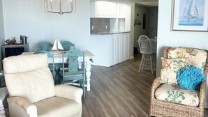 A living room with a beige armchair, a wicker chair with a blue pillow, a dining table with chairs, a kitchen area, and a framed picture on the wall.
