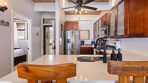 A compact kitchen with wooden cabinets, a stainless steel refrigerator, a built-in oven, and a ceiling fan. - Oceanfront, remodeled penthouse, sunsets, whales/dolphins, surfers! Free parking (Kailua-Kona)