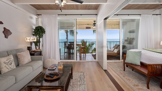 A spacious living area with a sofa, coffee table, and a view of the ocean through large sliding glass doors. - Oceanfront, remodeled penthouse, sunsets, whales/dolphins, surfers! Free parking (Kailua-Kona)