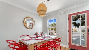 Un coin repas avec une table blanche dressée pour quatre personnes, des chaises rouges, un miroir rond et un luminaire suspendu en osier.
