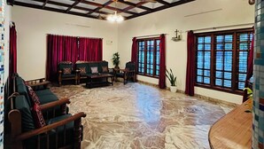 Una habitación amplia con techo alto, vigas de madera, piso de mármol y ventanas grandes con cortinas rojas.