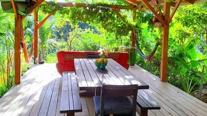 An outdoor wooden deck with a table and benches, surrounded by lush greenery and a hanging bunch of bananas. - Secluded Mountain Retreat w/ river @ Finca Remedio Eco Farm (Utuado)