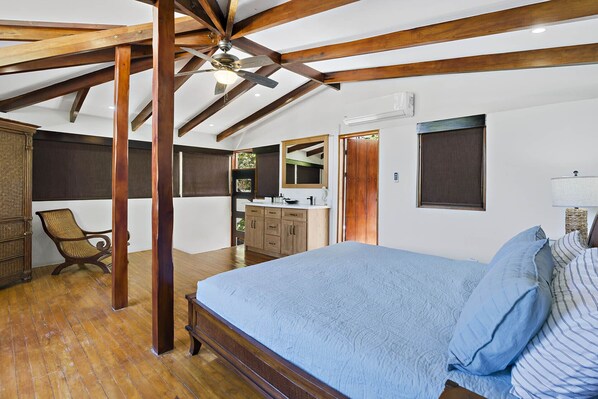 A bedroom with a large bed, wooden beams, a ceiling fan, and a view of the kitchen.