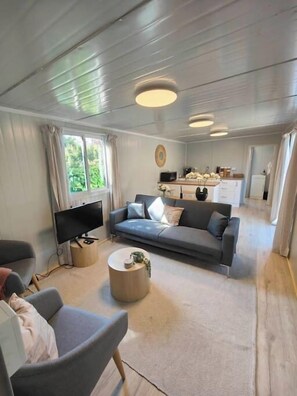 A modern living room with a grey sofa, a TV, and a coffee table. There is a kitchen in the background with cabinets and a refrigerator. - 30m² Tiny Home w/Parking | 5 mins to One NZ Stadium (Christchurch)