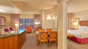 A hotel room with a bed, a small table, a desk with a chair, a dining area with chairs, a window with curtains, and a bathroom with a sink and mirror. - Las Vegas | Vegas Weekend Escape | Pool & Resort | 2BR (Las Vegas)
