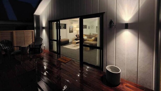 Una casa moderna con una terrazza in legno, un'ampia vetrata che dà su un soggiorno e un lampadario a parete.