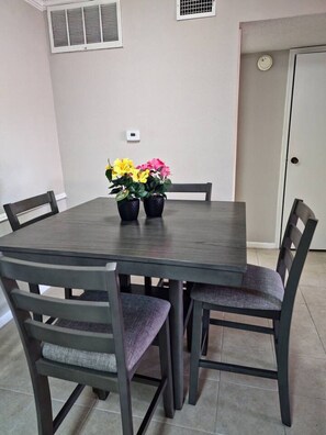 A dining area with a dark wooden table and four chairs, each with a cushioned seat. There are two potted plants with yellow and pink flowers on the table. - A Private Retreat Made for Extended Houston Stays (Houston)