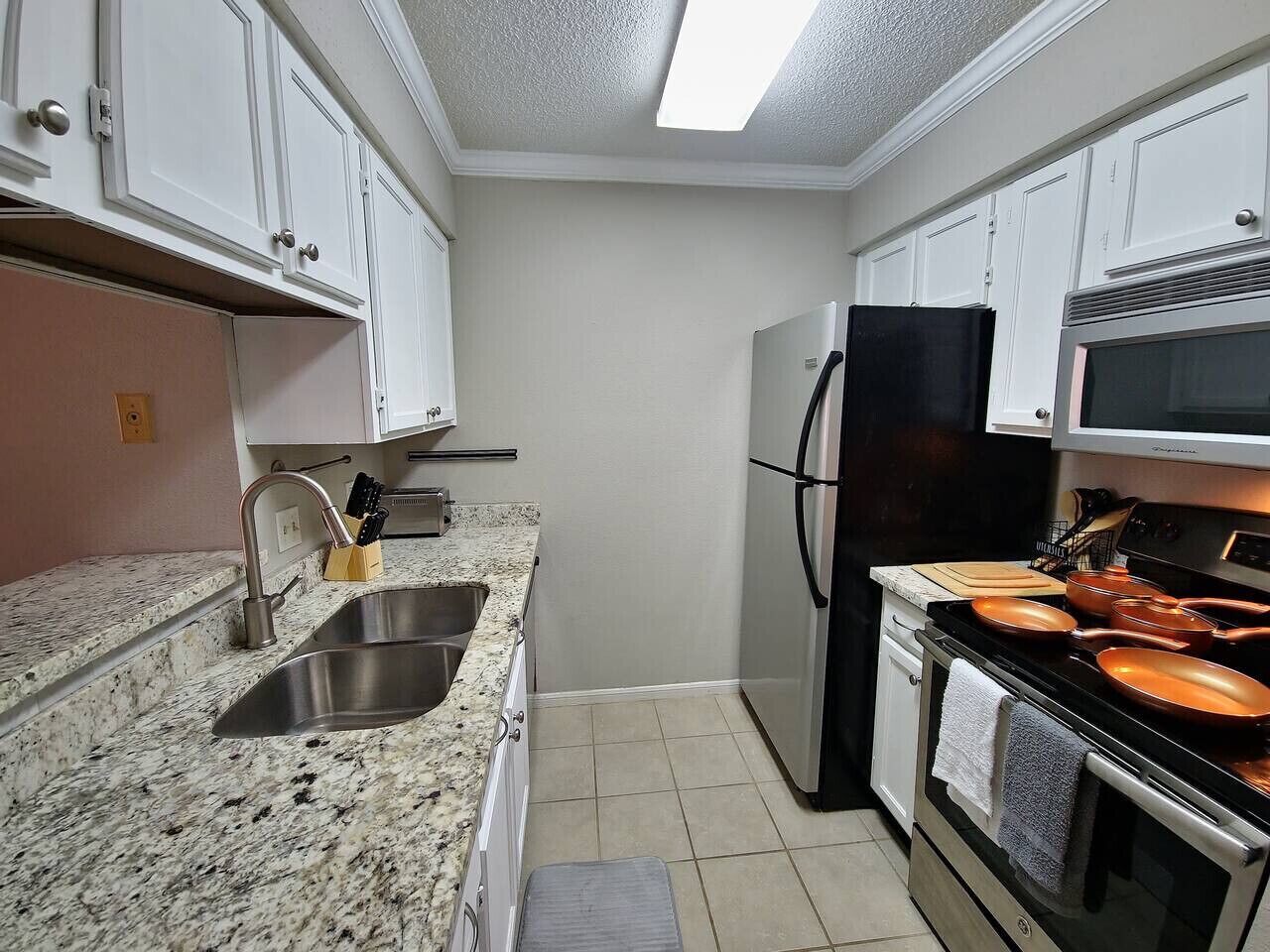 Una cocina compacta con armarios blancos, un lavamanos de acero inoxidable, un refrigerador negro y una estufa con ollas de cobre.