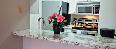 A kitchen with a granite countertop, a sink, a potted plant, and a microwave. There are two bar stools in front of the counter.