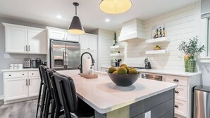 Una cocina moderna con armarios blancos, una isla central con un frutero, taburetes negros y lámparas colgantes.