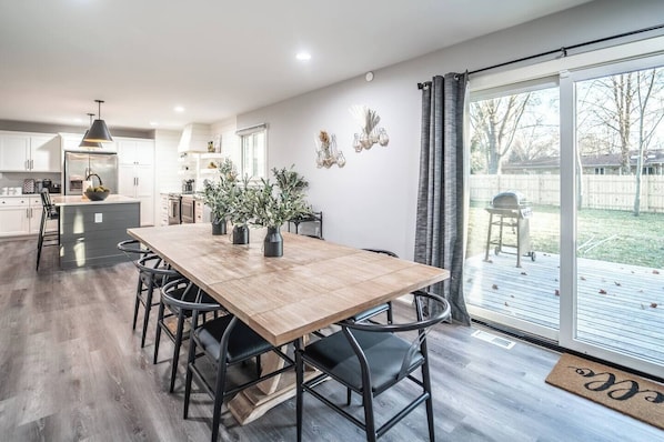A modern kitchen with white cabinets, a wooden dining table, and black chairs. There is a large window with a view of the backyard. - OliveBranch-LuxeDesigner Home-Spa-Firepit-GameZone (Greenwood)