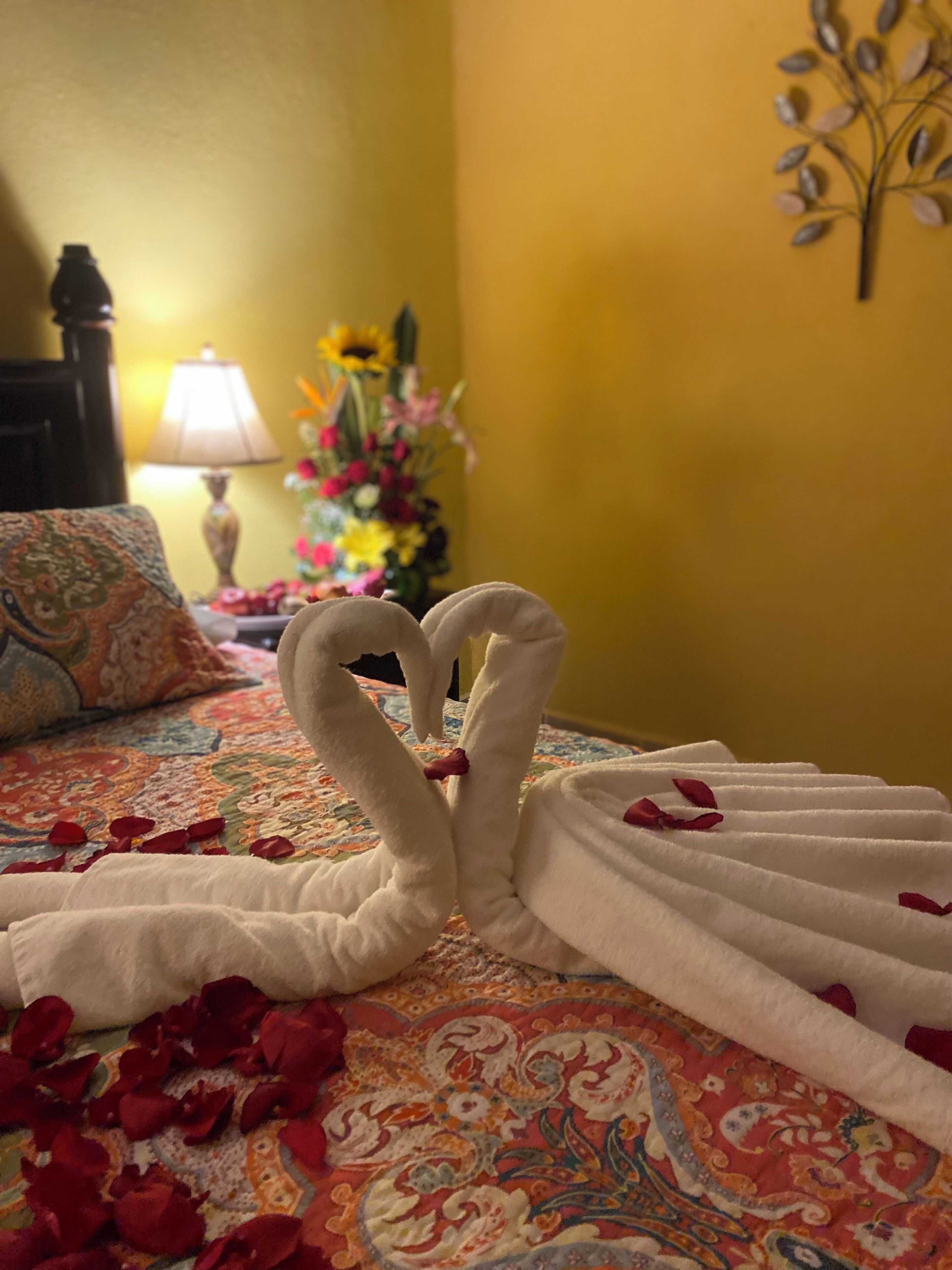 A bed with a decorative towel arrangement in the shape of swans, rose petals scattered on the bed, and a floral arrangement in the background.