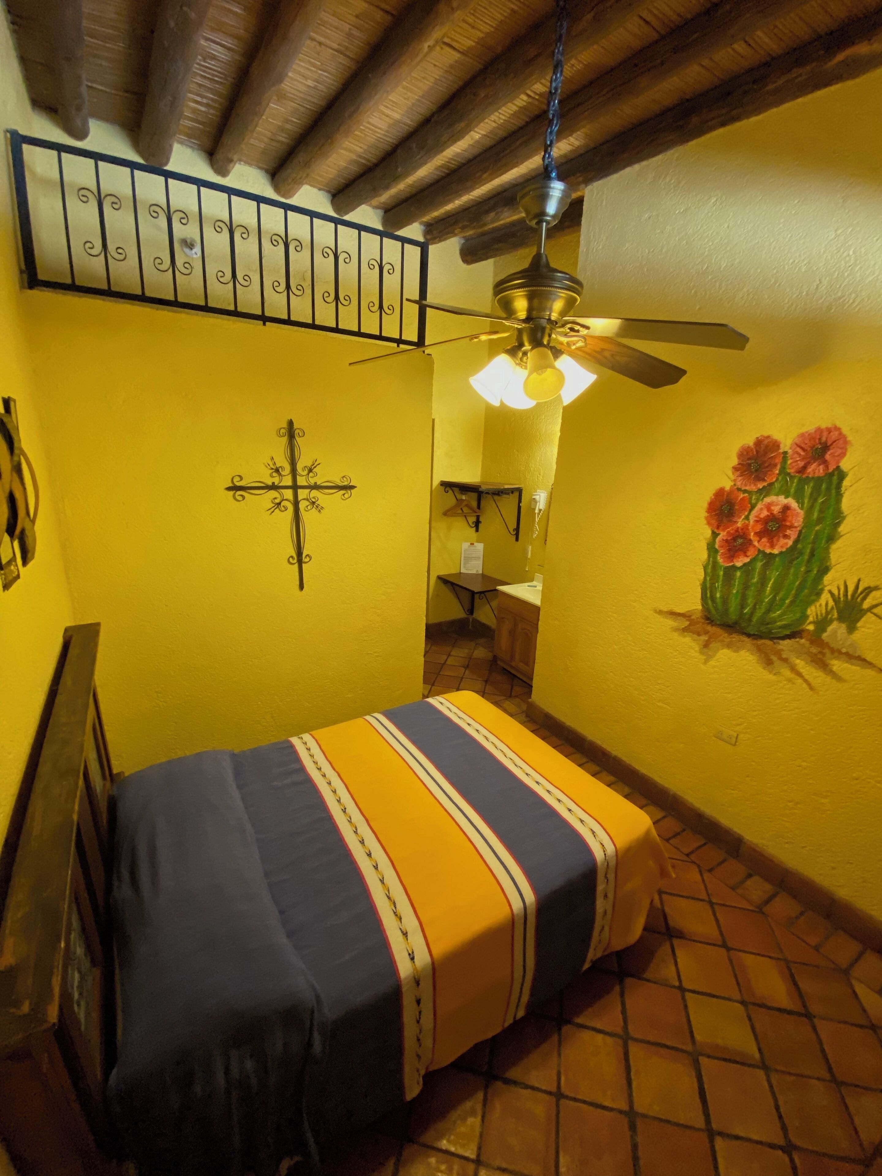 Una habitación con una cama, un ventilador de techo, un cactus pintado en la pared y una cruz en la pared.