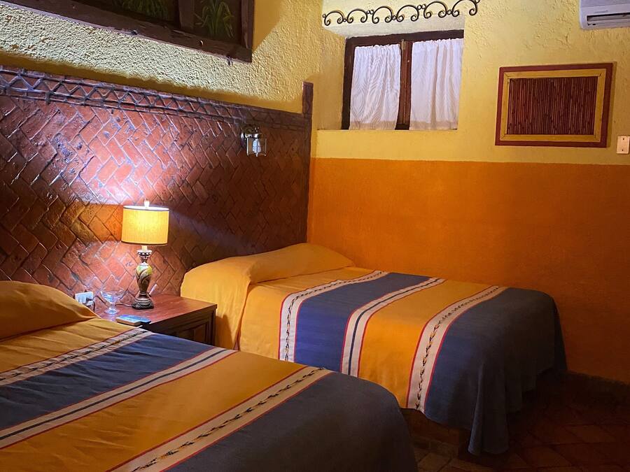 Una habitación con dos camas, una mesita de noche con lámpara, una ventana con cortinas y un panel decorativo en la pared.