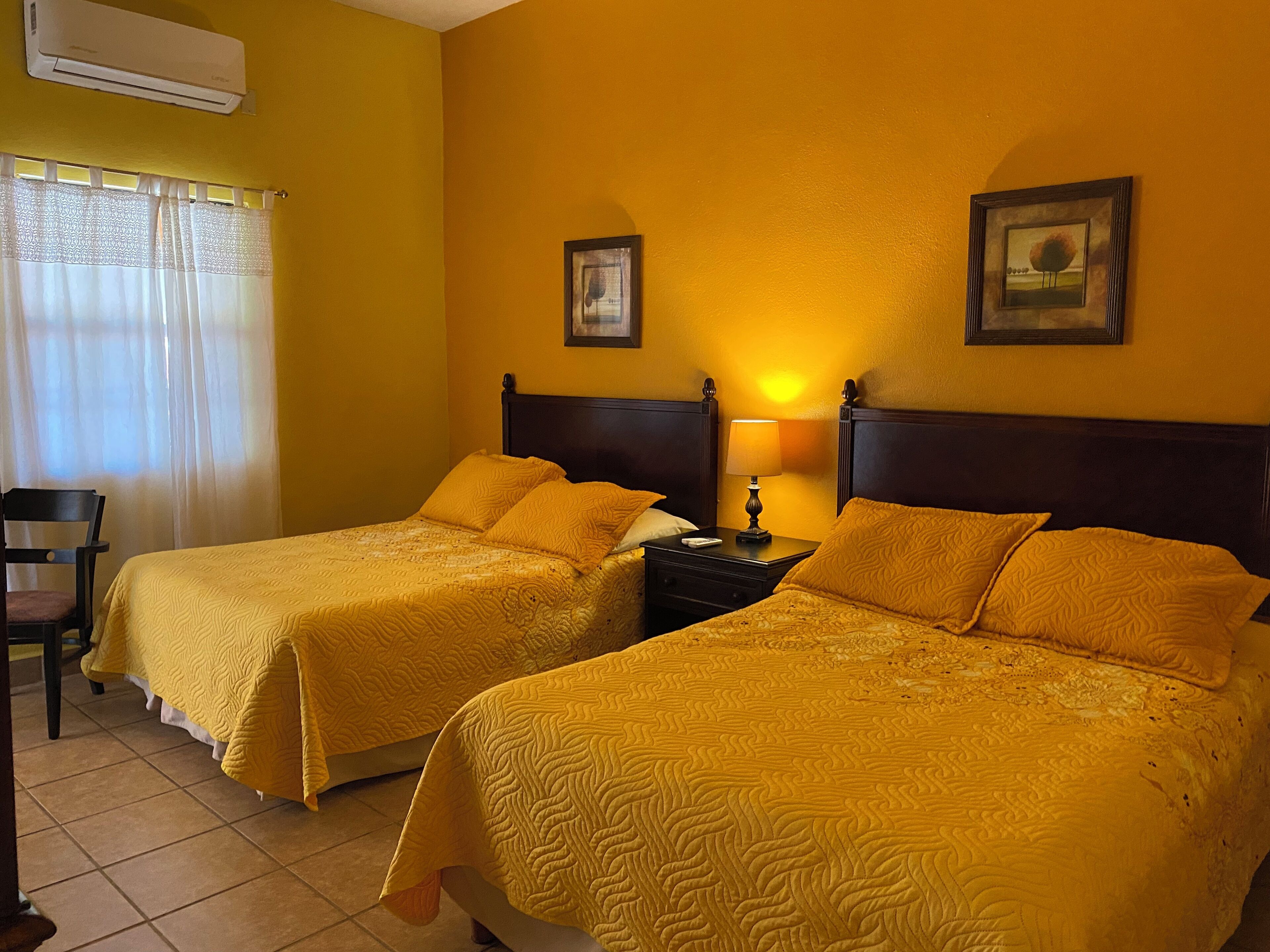 A hotel room with two beds, a chair, a nightstand with a lamp, and two framed pictures on the wall.