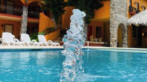 A swimming pool with a water feature in the foreground, surrounded by lounge chairs and a building with a red roof and stone pillars. - Hotel Quinta Sta Cecilia (Cuatro Ciénegas)