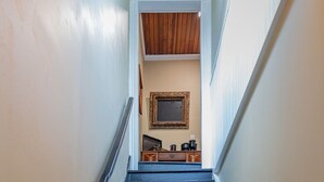 Uma escadaria com degraus de madeira escura e paredes brancas, que leva a um quarto com teto de madeira e um quadro na parede.