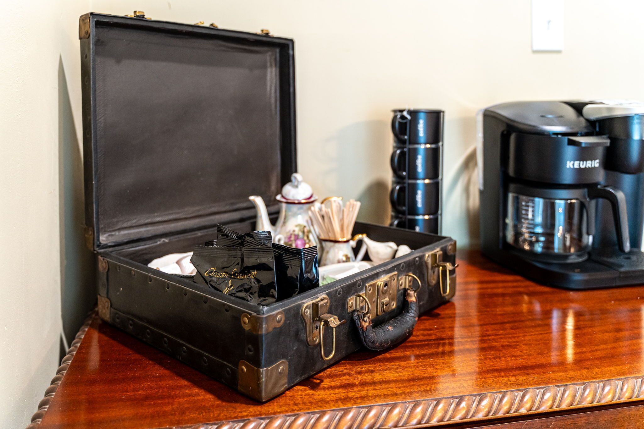 A vintage-style black suitcase with brass accents on a wooden table, containing tea bags, a teapot, and a cup.