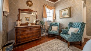 Quarto com cômoda de madeira, poltrona azul, tapete estampado e espelho antigo.