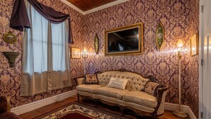A room with a patterned wallpaper, a tufted sofa, a floor lamp, and a large window with curtains. - TheOutPostInn Bed&Breakfast - The Purple Victorian (Marthasville)